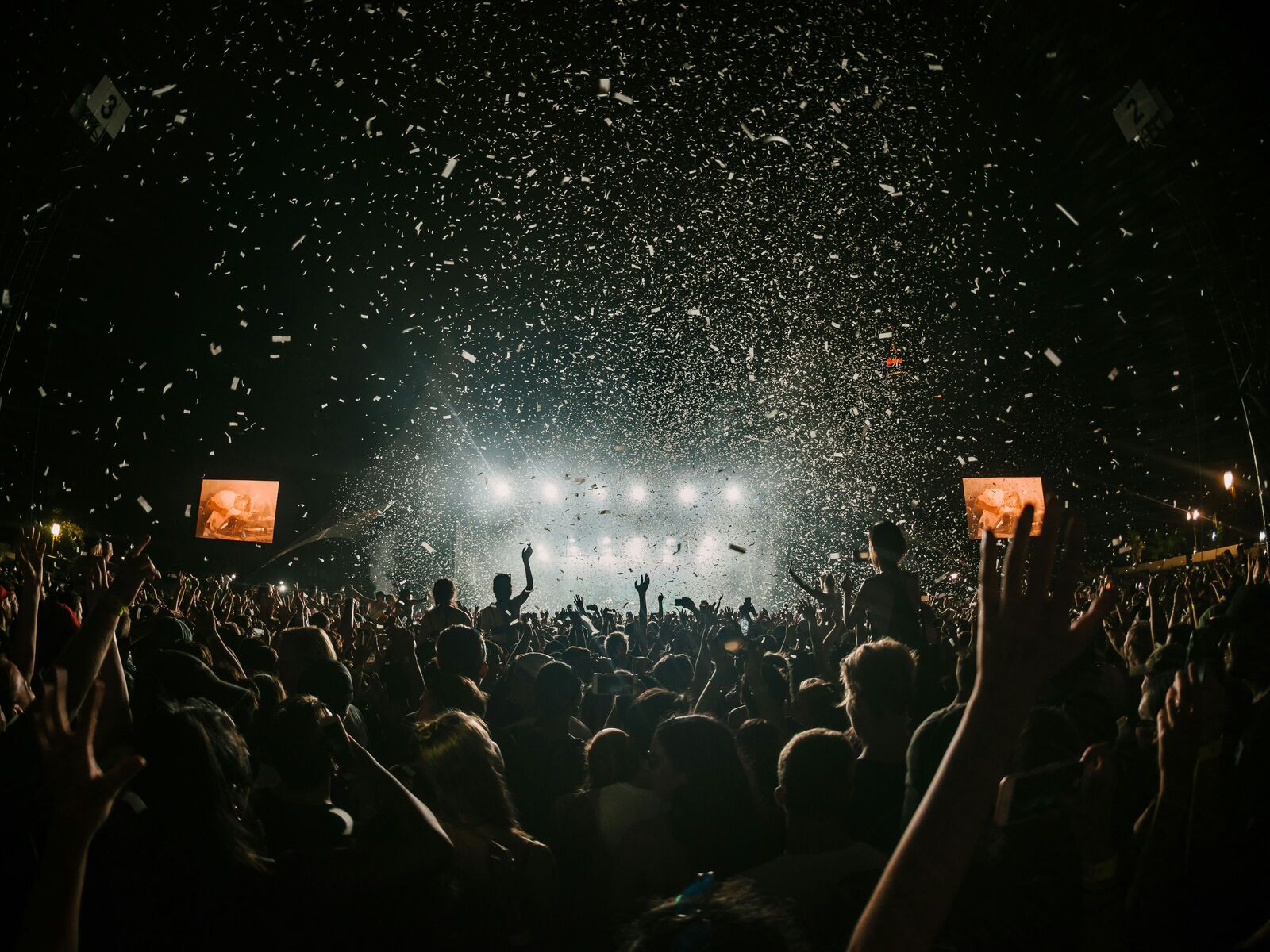 A festival crowd with hands raised as confetti falls in front of a brightly lit stage